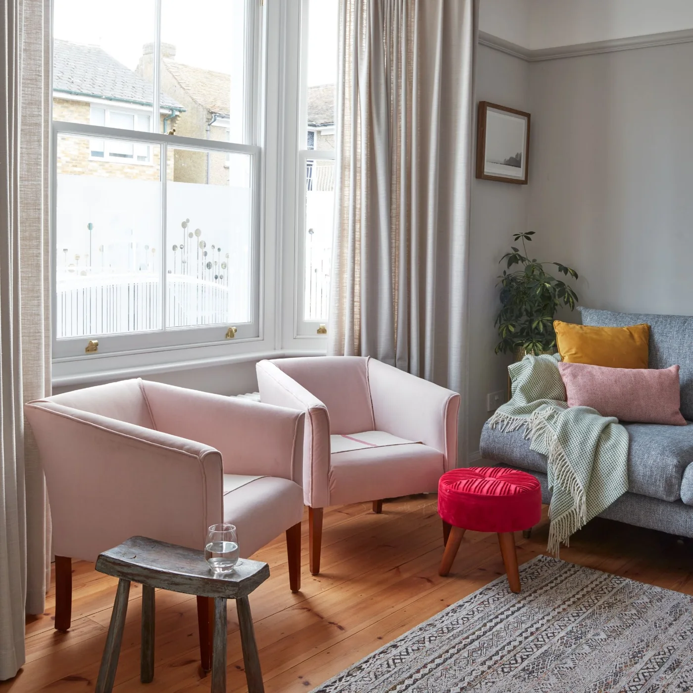 A gray sofa shares a bright sitting room with two pale pink chairs, a red stool, patterned rug and light curtains