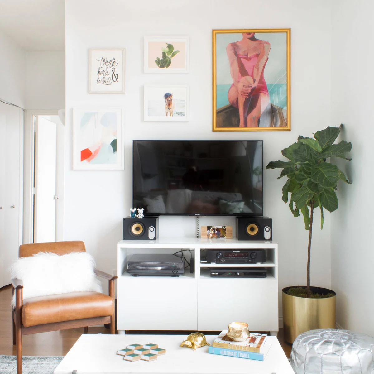 Pink figure painting and small pale-framed prints above a television on a white media cabinet beside a tall plant.