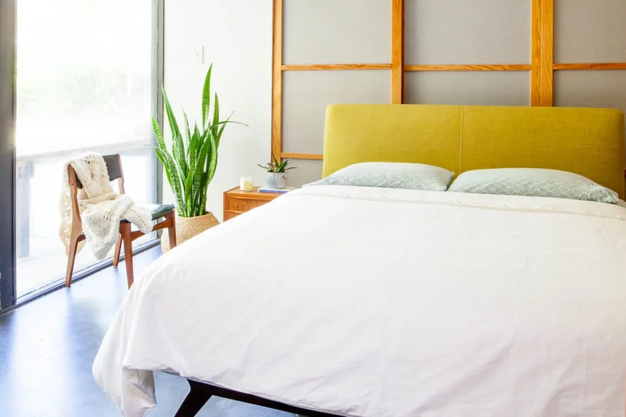 A rectangular yellow upholstered headboard sits against a wood grid with white bedding, a wood bedside cabinet, plants and a chair beside glazing.