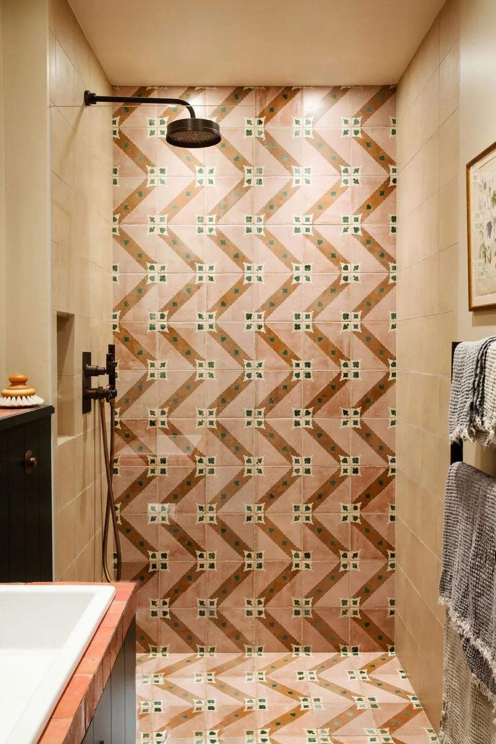 A shower with a clear side screen has pink, ochre and green patterned tiles across its back wall and floor, with dark fittings.