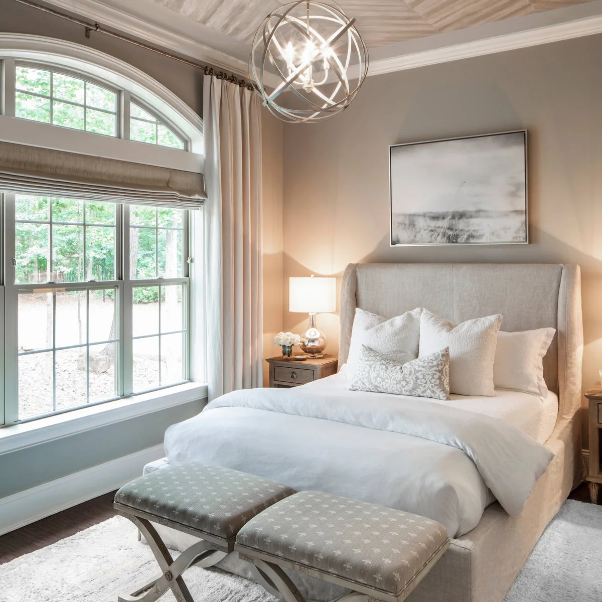 Soft gray landscape picture above a pale upholstered bed beside an arched window and long curtains.