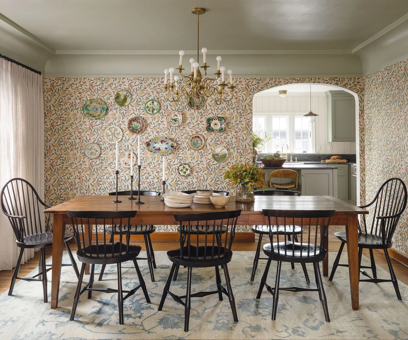 Dining room with botanical wallpaper, decorative wall plates, a wooden table, black spindle chairs and a gold-colored chandelier.