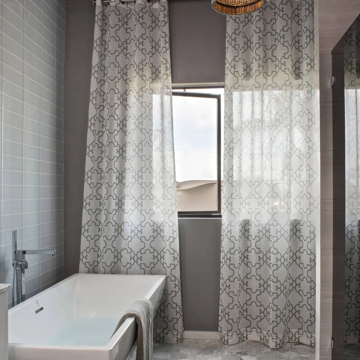 Tall sheer white panels with a gray outline pattern hang beside a bath and an open window.