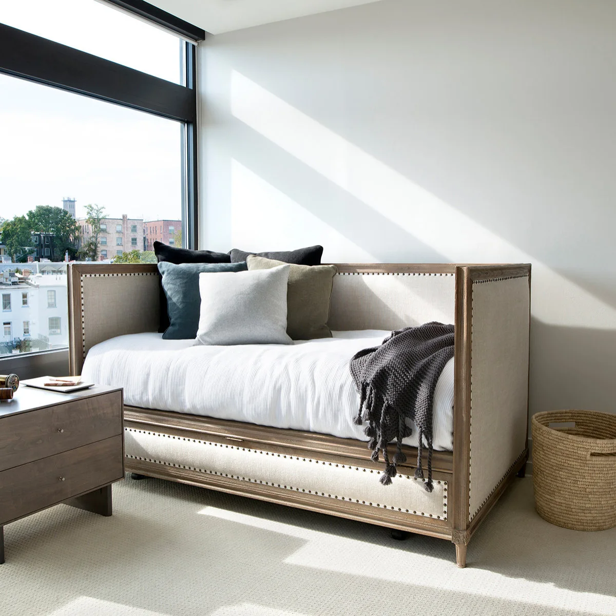 A pale upholstered daybed with a wood frame and nailhead trim stands beside a large window and a small chest.