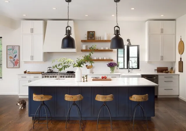 A blue island stands below black pendants in a pale kitchen with timber shelves.