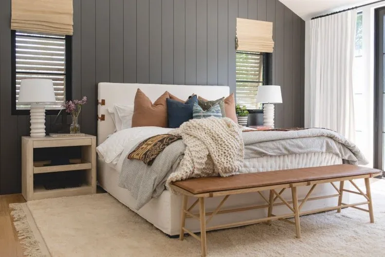 Charcoal vertical wall boards stand behind a pale upholstered bed, timber bedside units and a narrow brown bench.