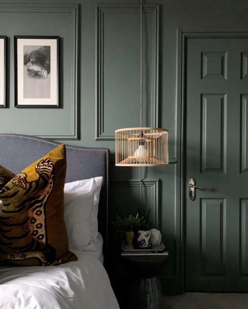 Dark green paneled bedroom with gray studded headboard, white bedding, yellow patterned cushion, woven pendant, and framed photographs.