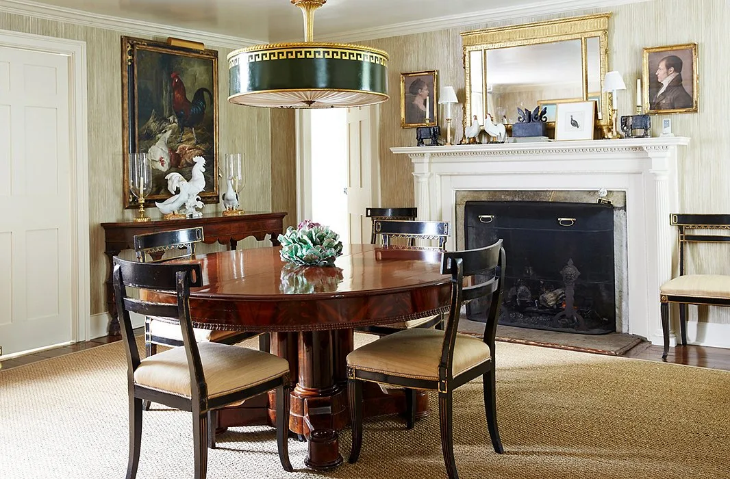 Round wooden pedestal dining table with dark chairs beside a white fireplace and gold-framed mirror.