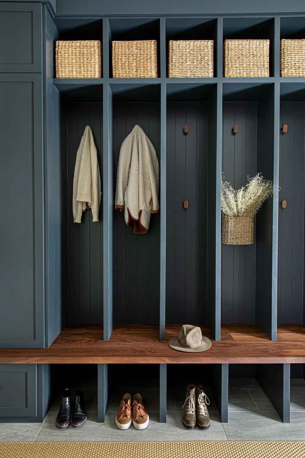 Tall deep navy mudroom lockers with woven upper baskets, coat hooks, wooden bench, lower shoe cubbies, side cupboard, gray floor, and pale woven rug.