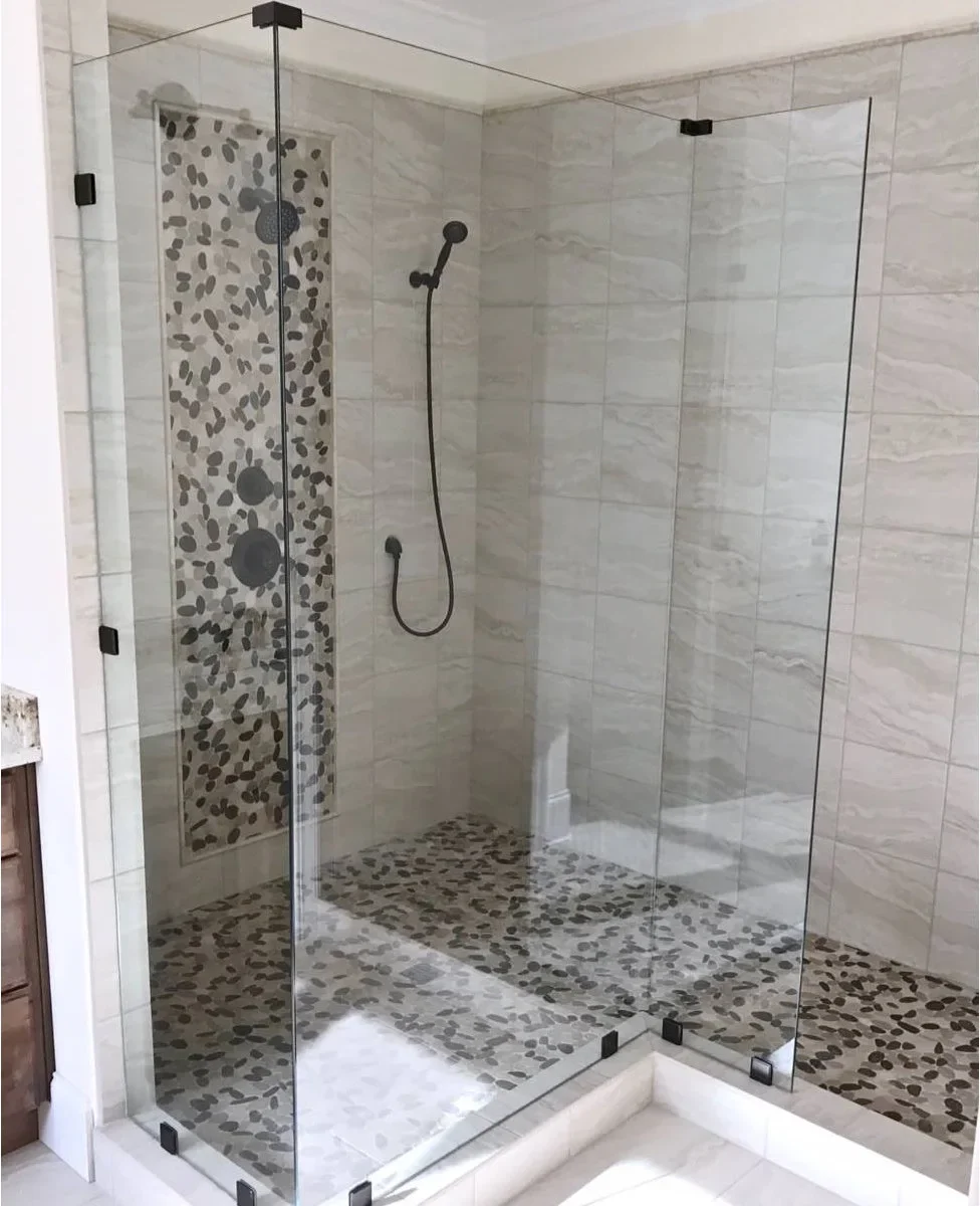 Clear fixed shower panels with dark clamps leave an open entrance beside a pebble-patterned floor and matching vertical tile strip.