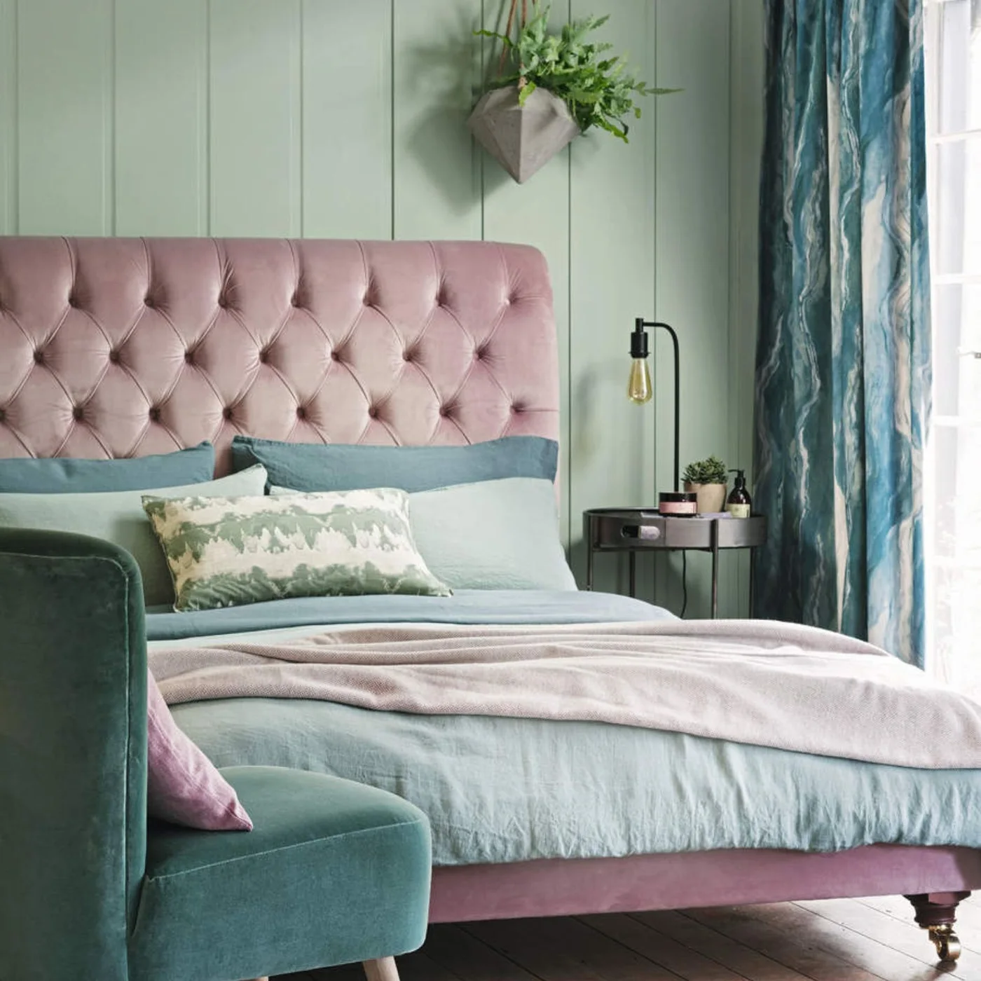A pink buttoned headboard stands beside pale green paneling, blue-green bedding and curtains with a small dark bedside table