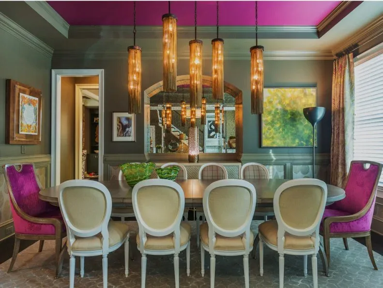 Pink upholstered end chairs with pale frames beside rows of pale oval-back chairs, a long dining table, gray walls, and hanging amber lights.