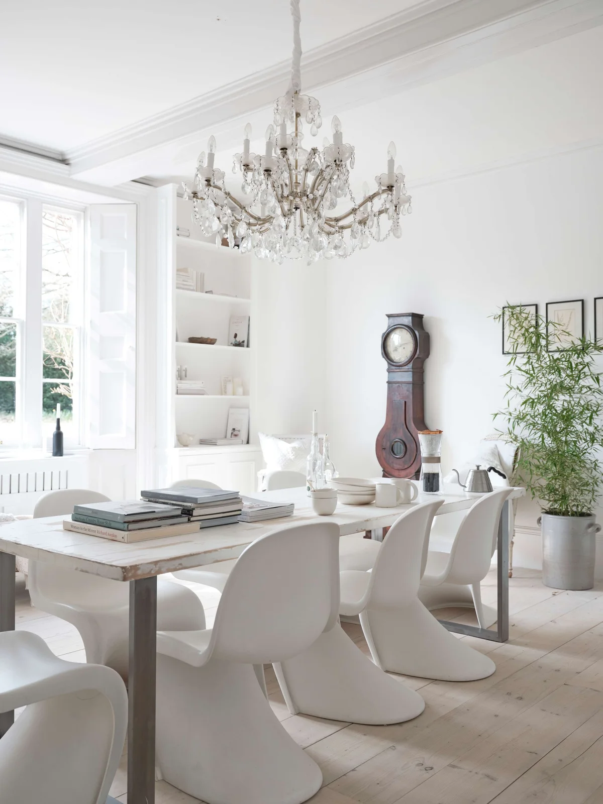 Curved pale cantilever dining chairs sit around a simple wood table beneath a decorative chandelier beside pale shelves, a tall wooden clock and light floorboards