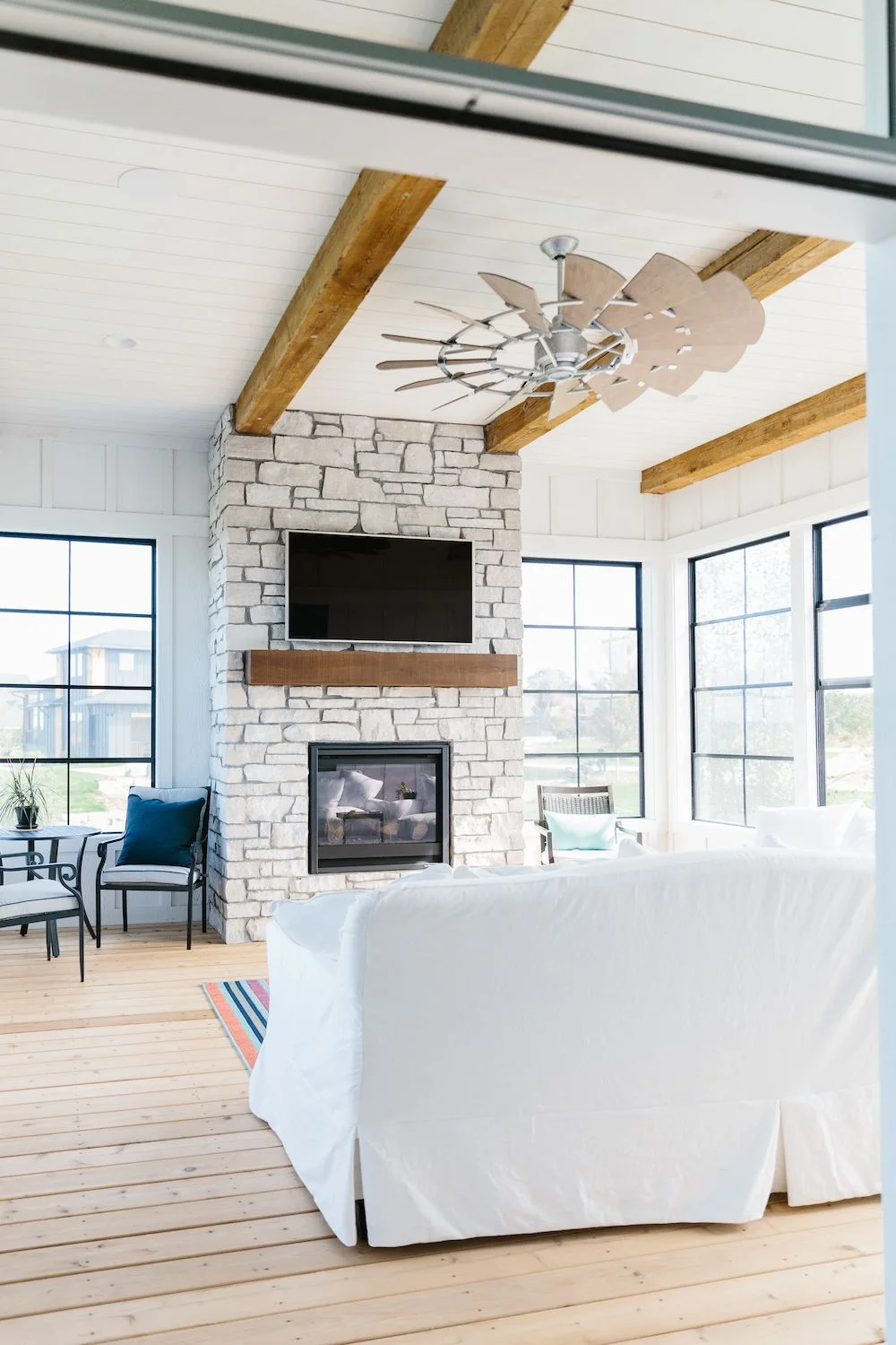 A pale stone chimney breast with a timber mantel and television stands among black-framed porch windows.