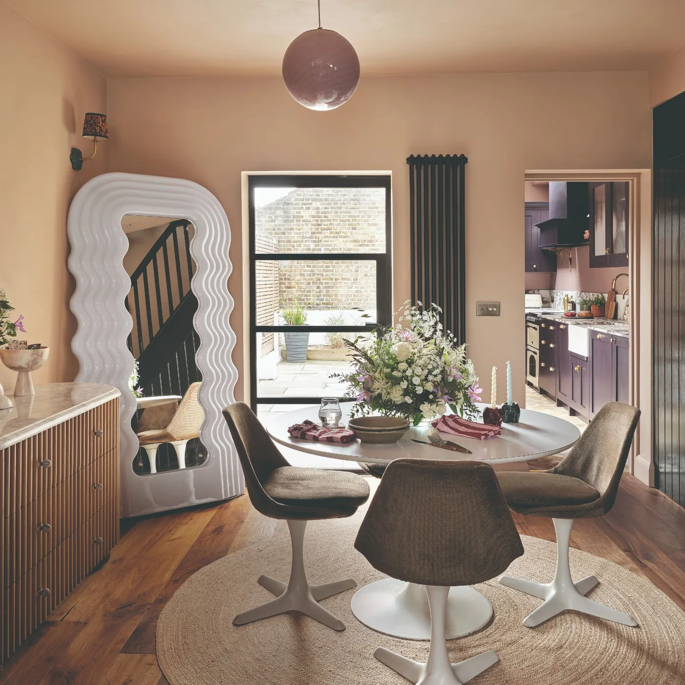 Blush dining room with a round white table, gray upholstered chairs, round woven rug, tall wavy mirror and wooden sideboard.