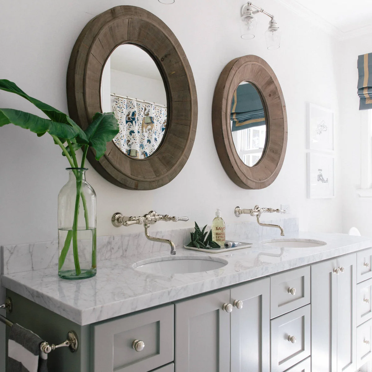 Light double vanity with a tall green bottle vase of broad leaves and a low tray of toiletries between the basins.