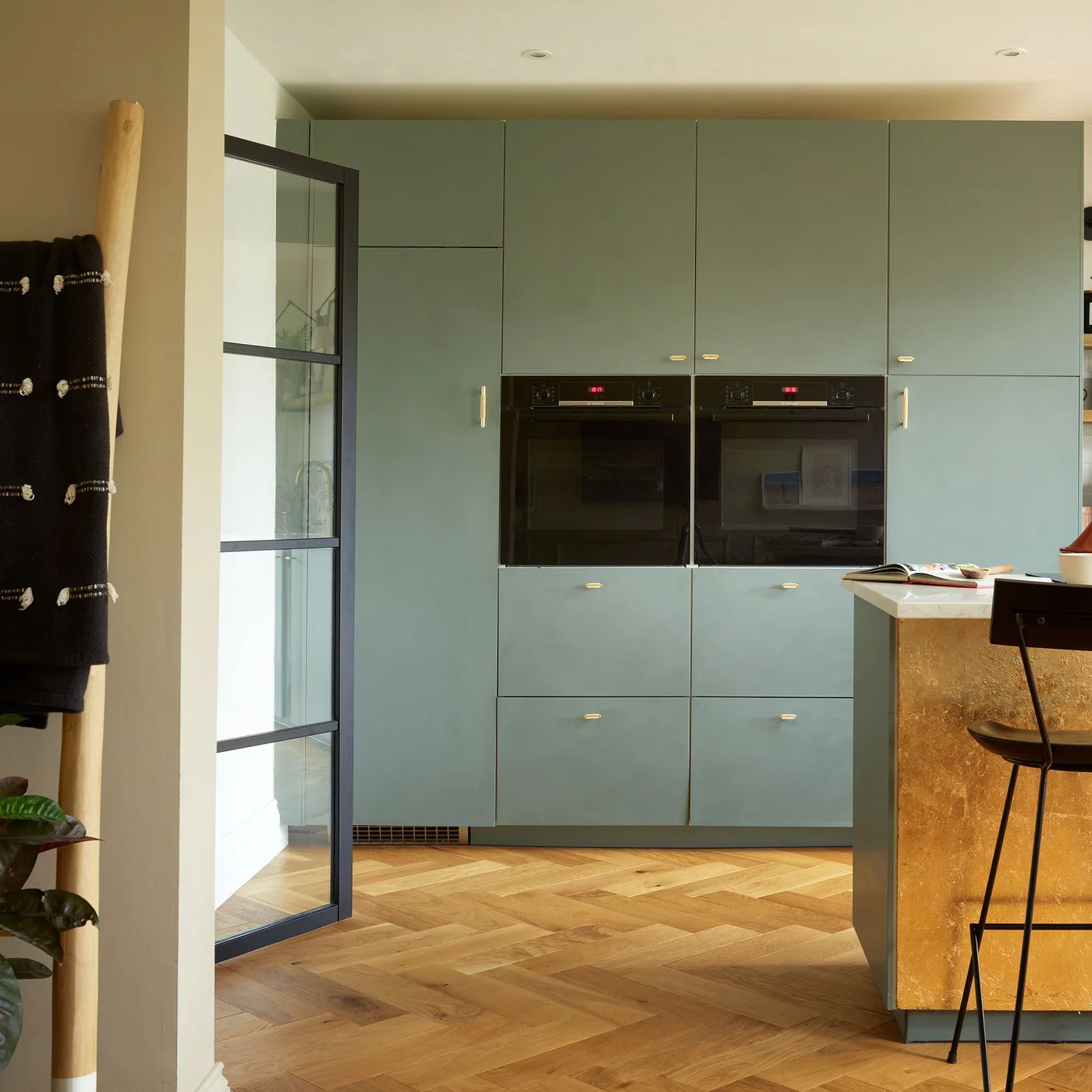 Muted green flat-front cabinets surround two black ovens beside a gold-colored island and a glazed door