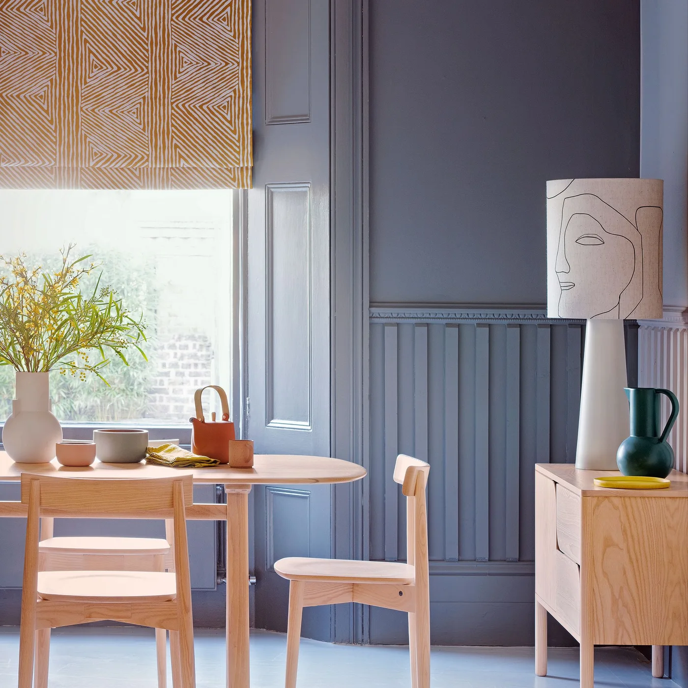 A large pale lamp with a line-drawn face and a dark green pitcher sit on a pale wood sideboard beside gray wall paneling.