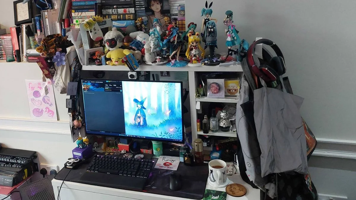 A compact white desk and hutch filled with books and character figures, with one monitor, headphones and a black keyboard below.