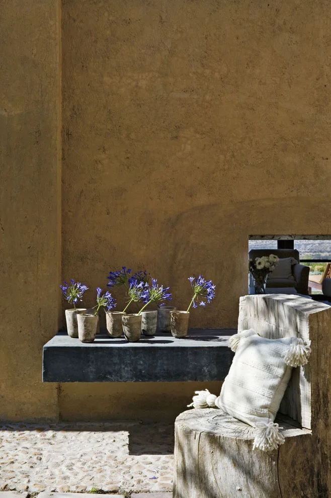 Small pots of purple flowers on a dark wall ledge beside a chunky wood chair with a pale tasseled cushion.