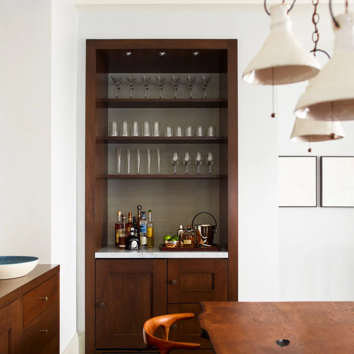 Wood-framed recessed mini bar with open glass shelves, bottle groupings and closed lower cabinets beside a dining table