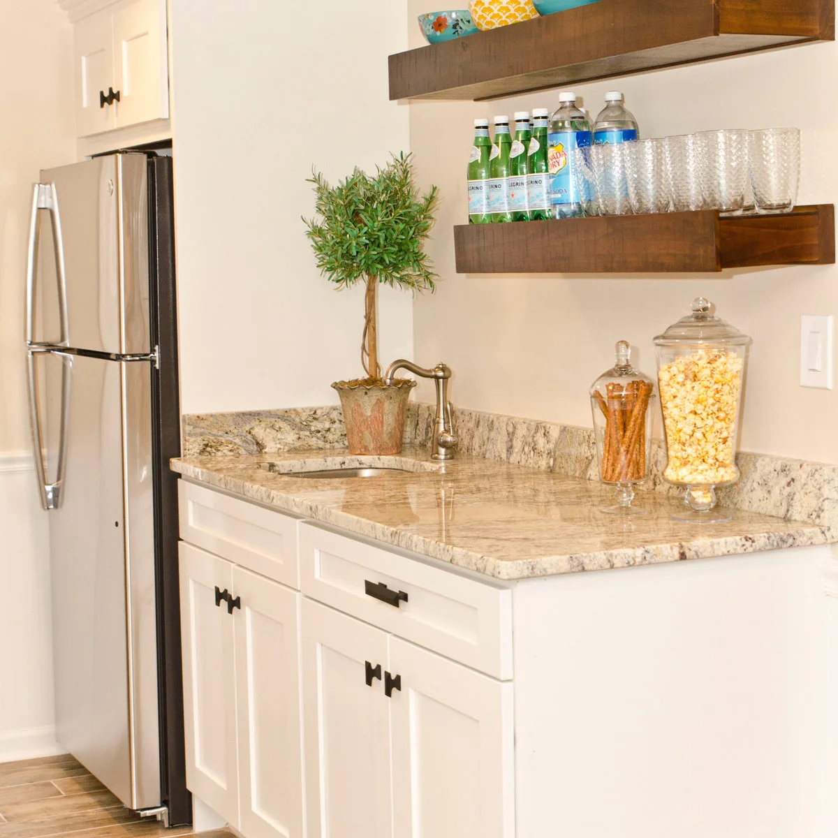 White sink cabinet beside a tall refrigerator with two wood shelves holding glasses and bottles