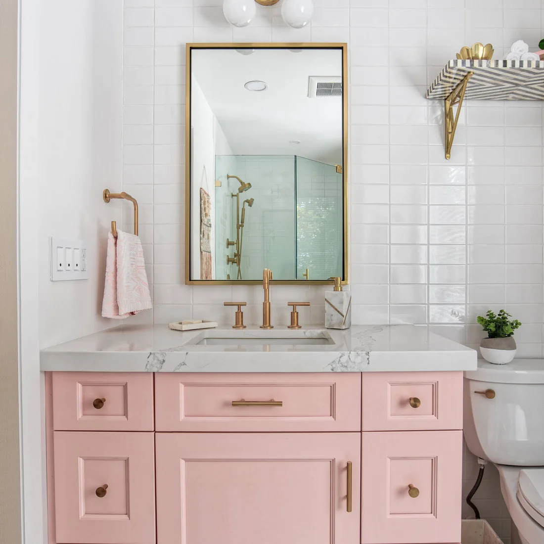 A pink vanity with warm metal handles sits below white tiles and a mirror reflecting a pale green shower.