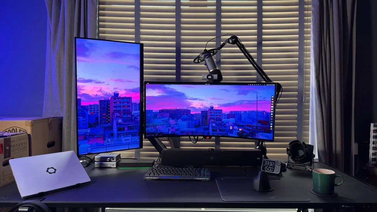 A dual-monitor desk in front of shutters, with a black microphone arm, a soundbar, a laptop and blue-purple accent light.