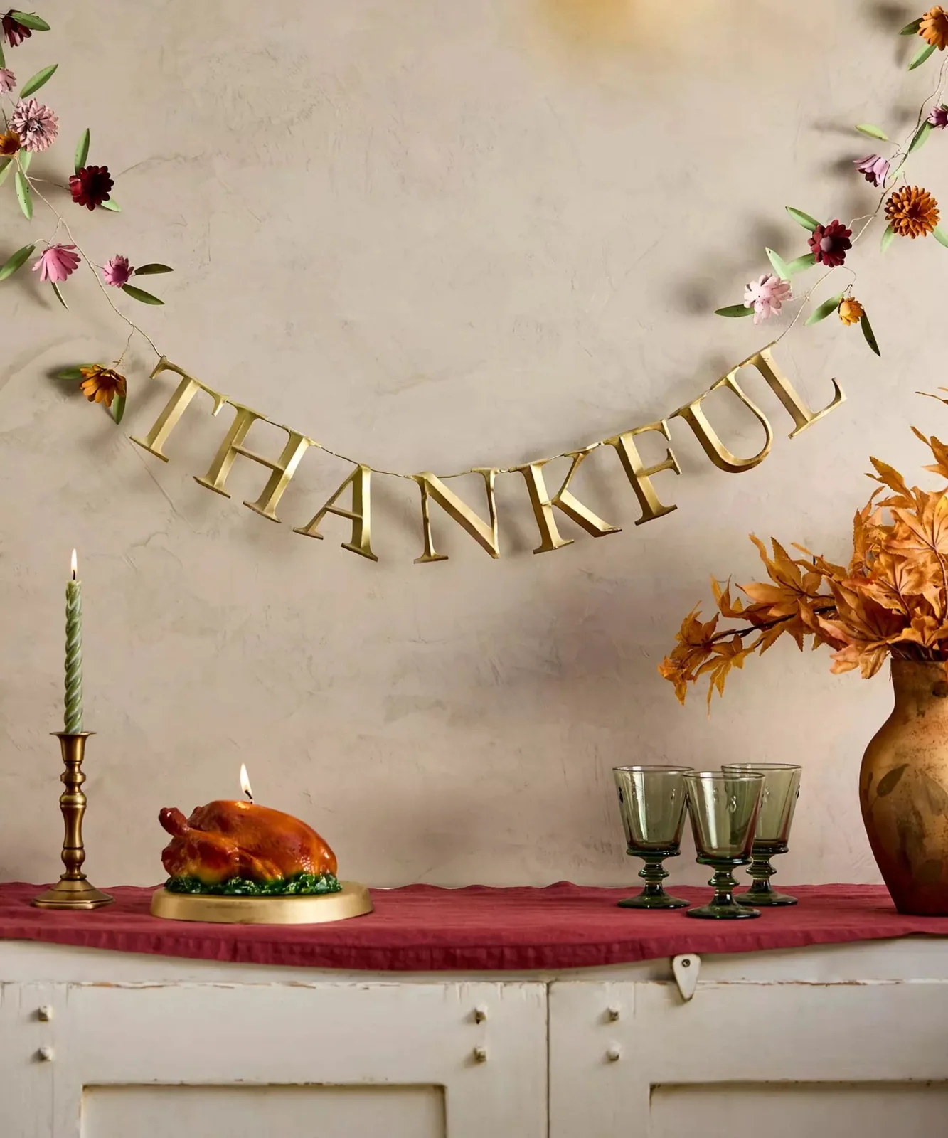 Warm metallic Thankful letters and small flower details hang above red cloth, green glasses and a turkey-shaped decoration