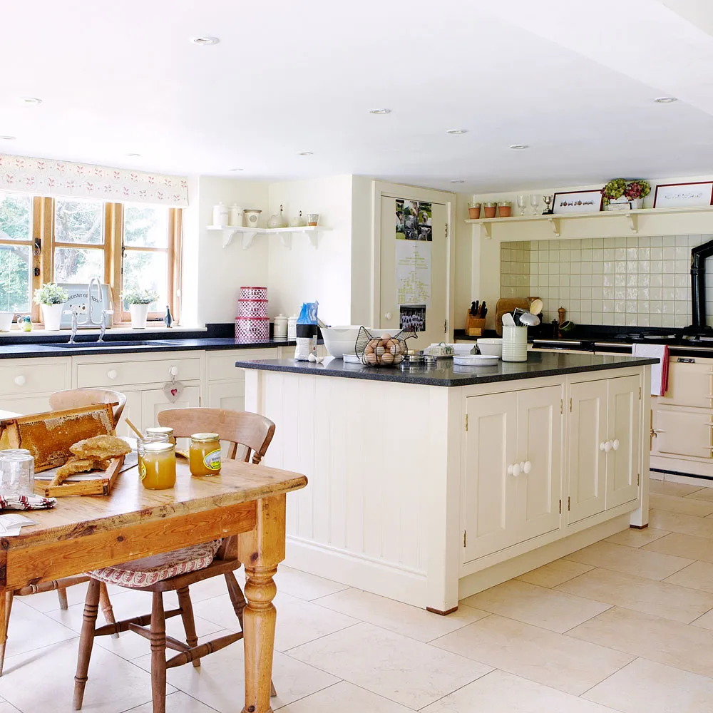 Cream-beige kitchen cabinets with dark counters, a pale island and timber dining table