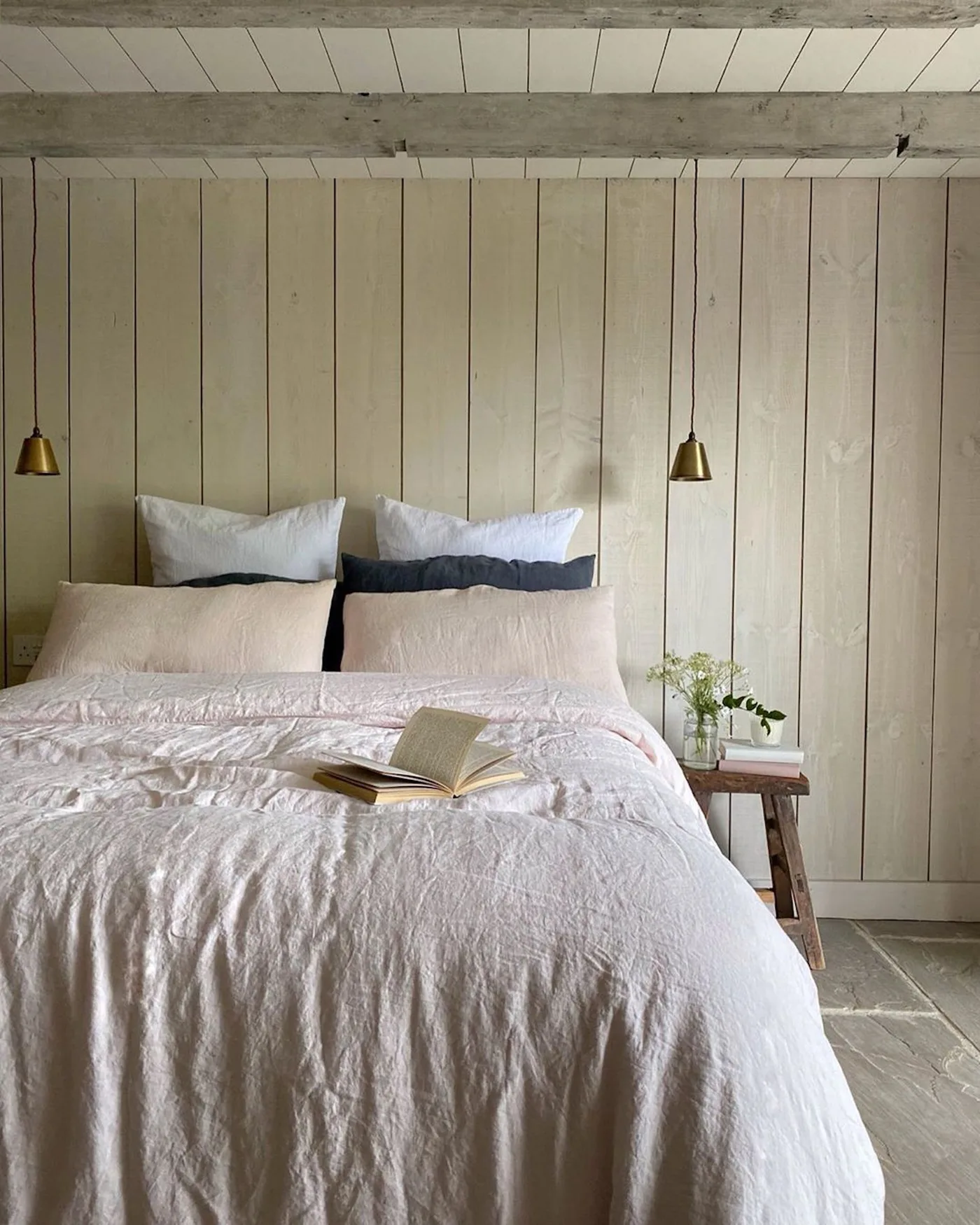 Pale vertical timber boards and ceiling beams frame a bed with soft neutral bedding and small hanging lights.