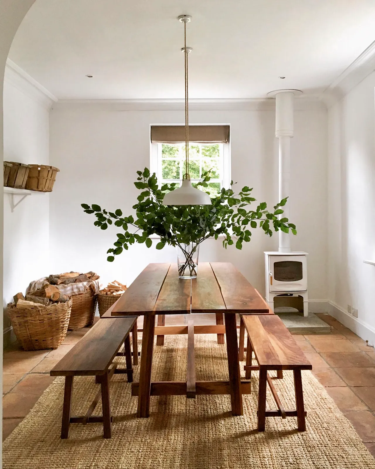 A wood plank dining table and two benches sit beneath a white pendant beside a white stove and baskets of logs.