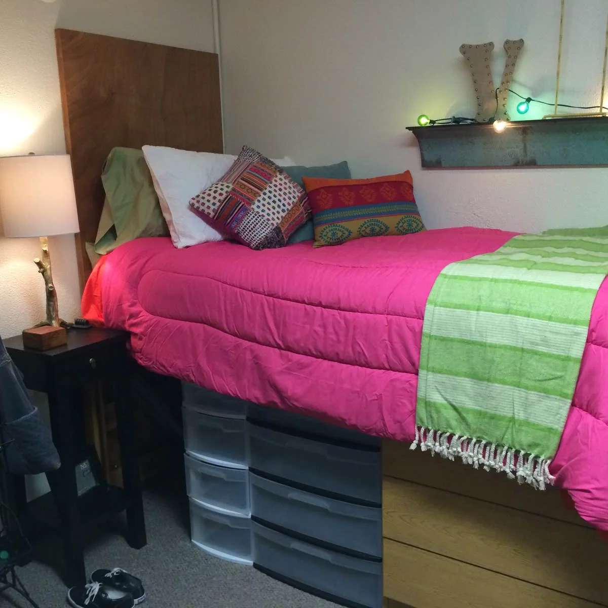 Raised wood dorm bed with pink bedding, green throw and plastic drawer units underneath