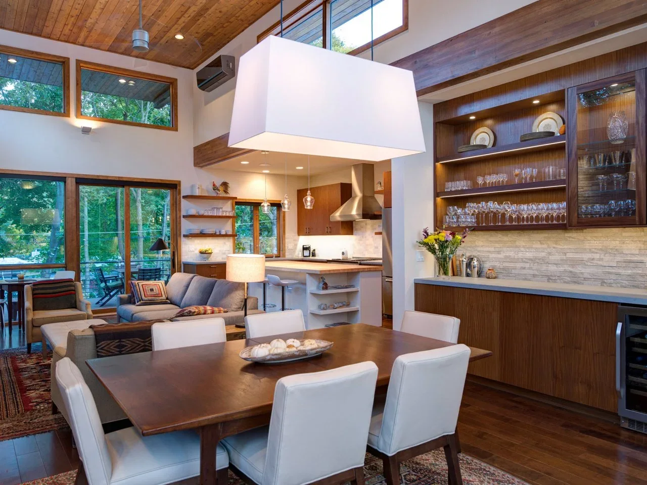A timber dining table sits beside glassware storage, an open kitchen and a small sitting area below a tall timber ceiling.