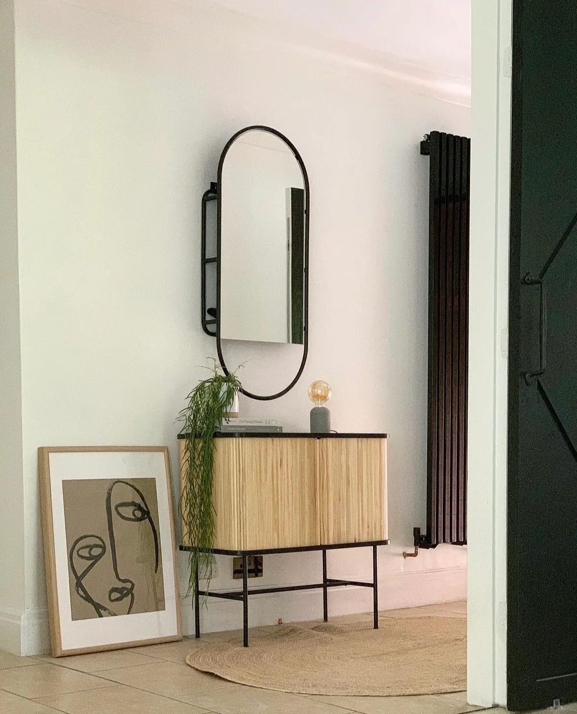 A small reeded timber cabinet on dark legs sits beneath a tall oval mirror beside a black radiator and trailing plant.