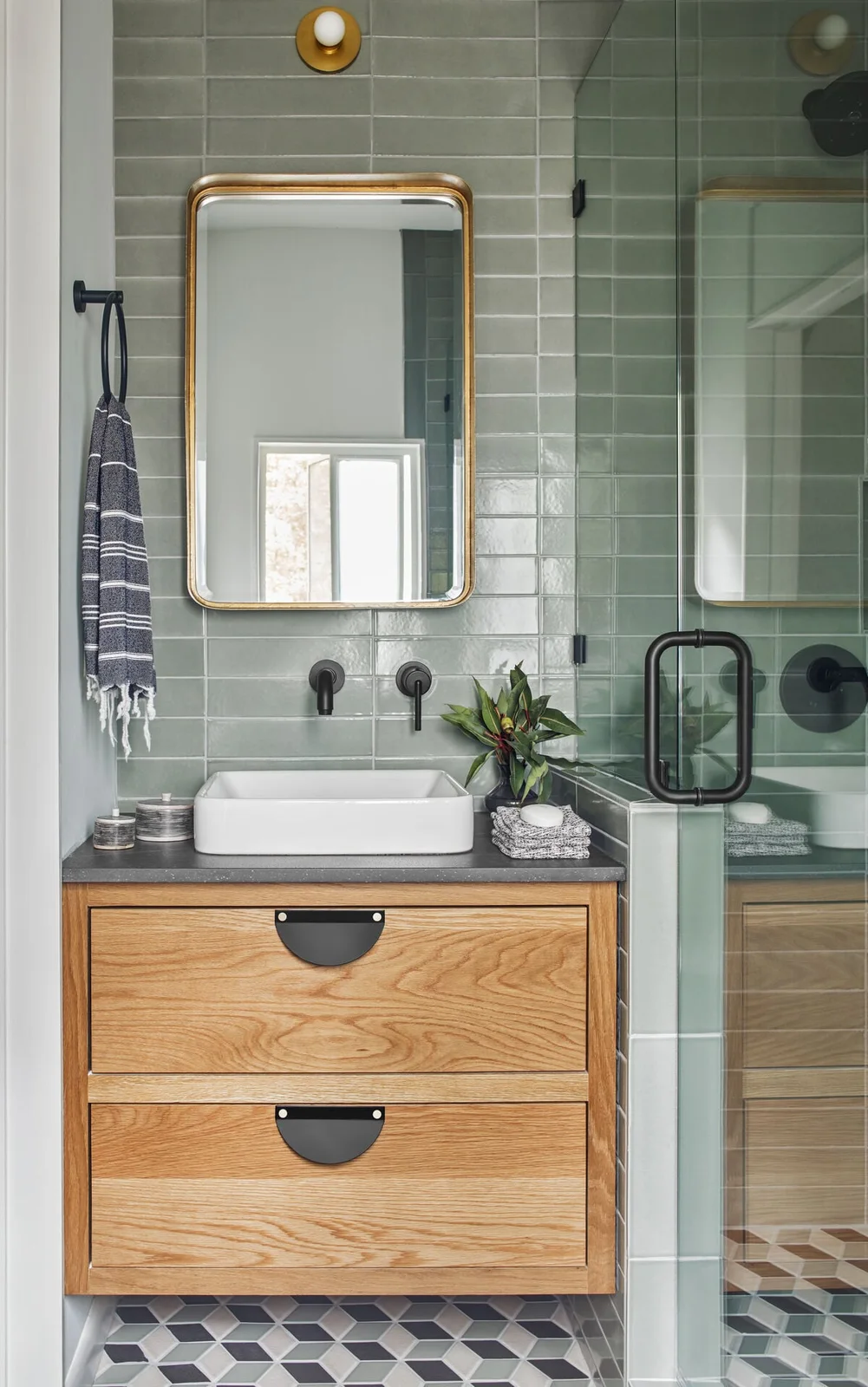 A compact wood drawer vanity with a white vessel basin sits against glossy sage tile beside a glass shower and a geometric floor.