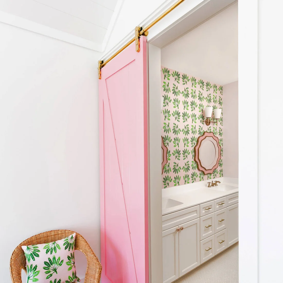 A pink sliding door frames a white vanity with green leaf wallpaper and a shaped pink mirror.