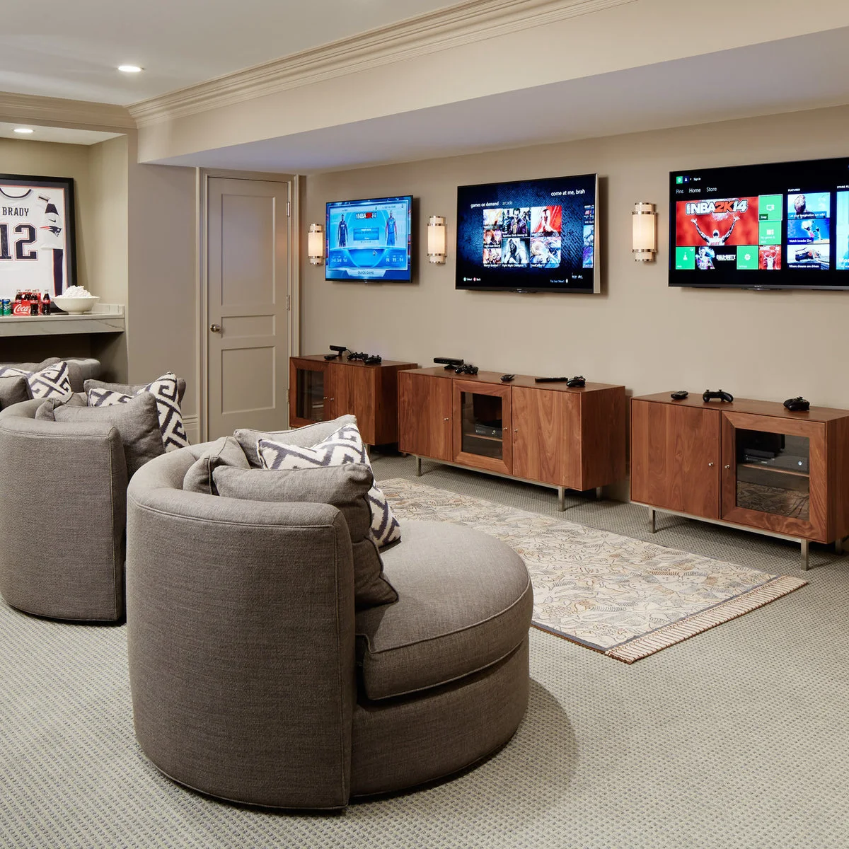 A room with three wall-mounted televisions, three gray rounded chairs, a long wood cabinet and dark painted walls.