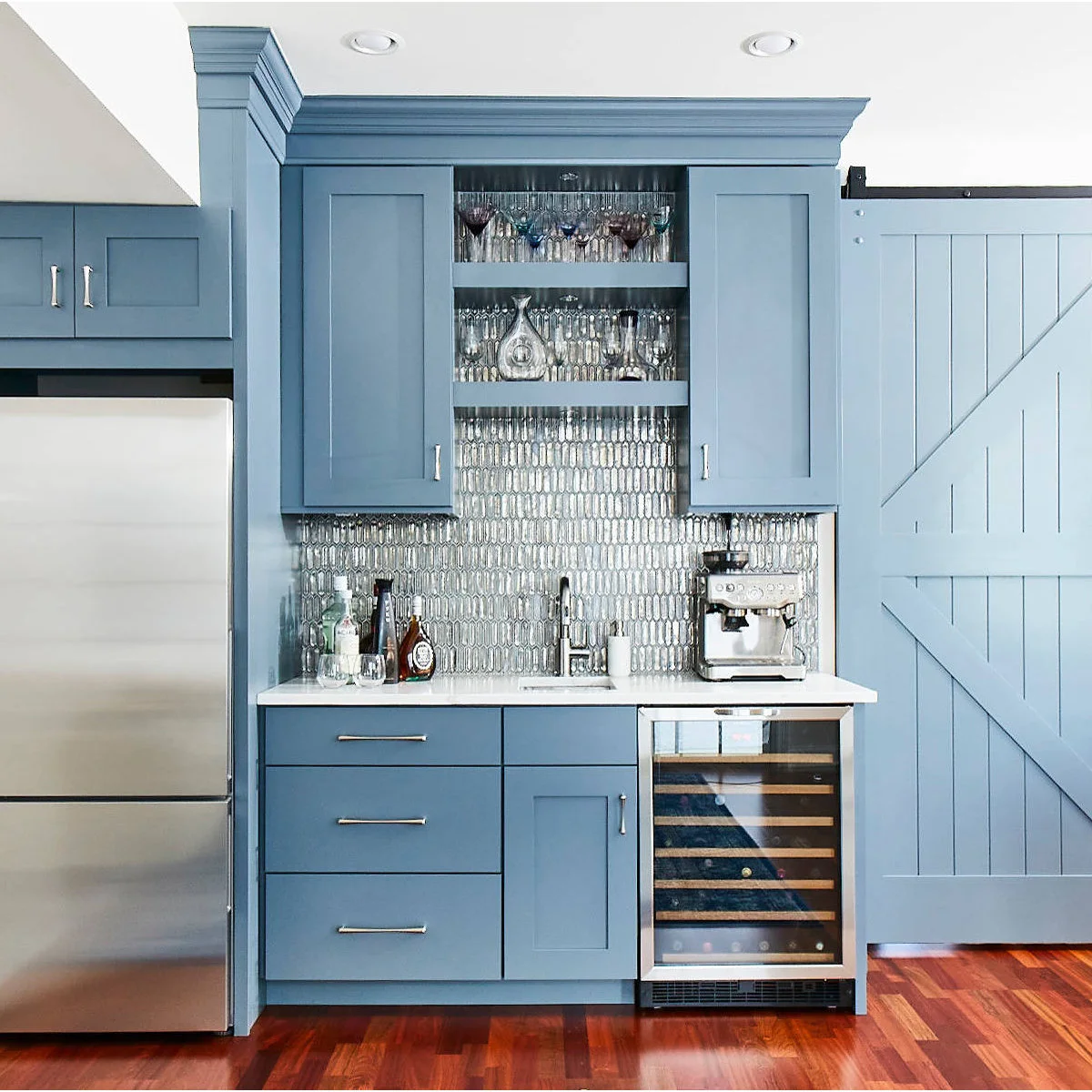 Blue cupboard doors above a stainless refrigerator match the blue cabinetry of an adjoining drinks station.