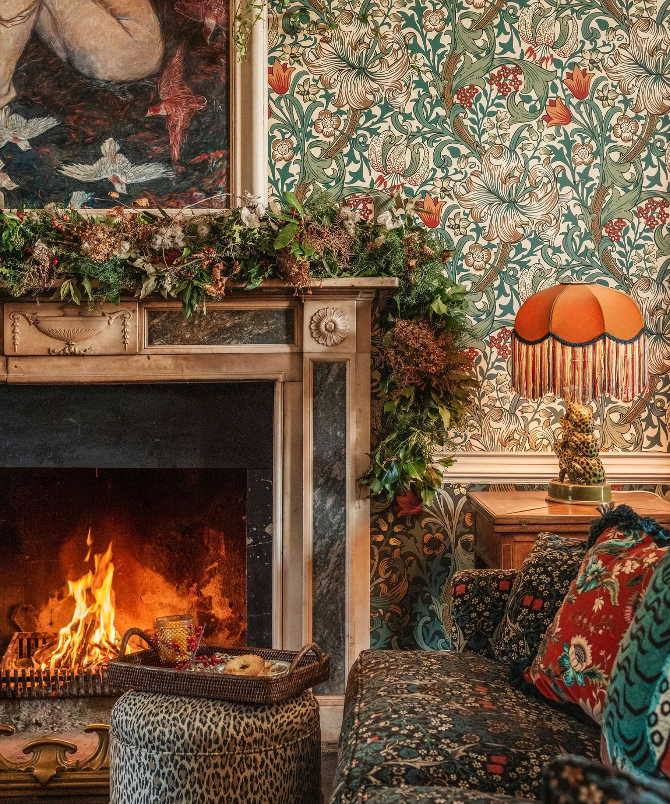 A green and brown foliage garland runs along a carved mantel beside patterned wallpaper, a warm-colored lamp and seating