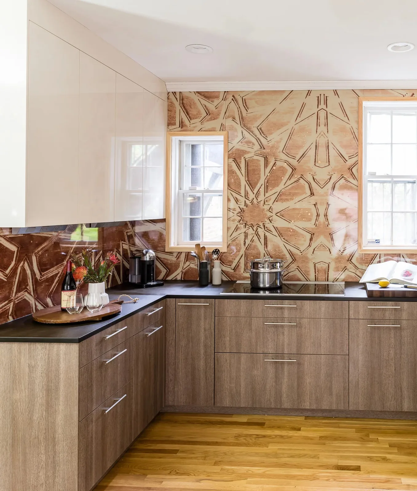 Plain wood-toned lower cabinets and white upper cabinets surround a black counter beneath a large warm-colored star pattern around two windows