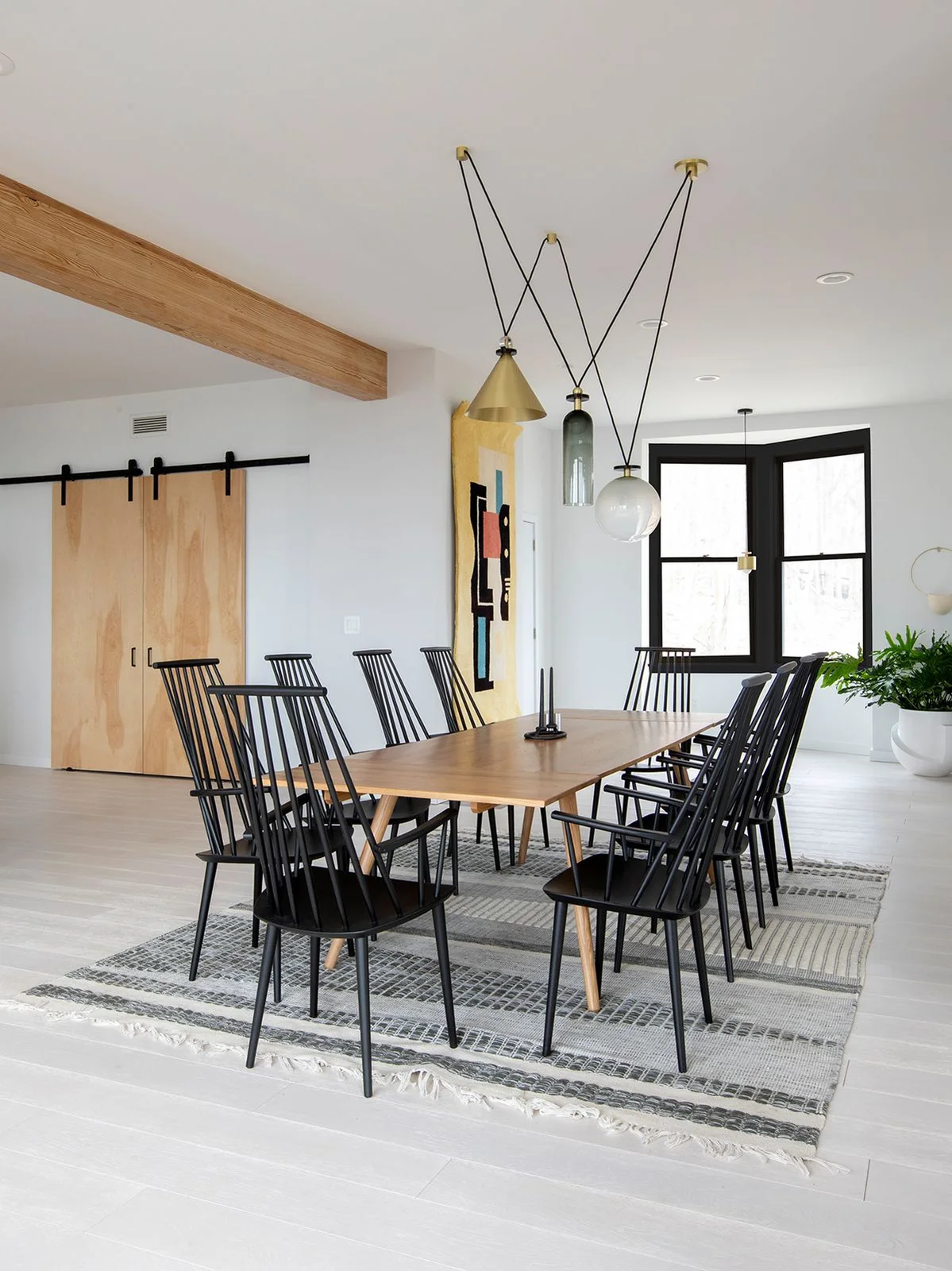 Black spindle-back dining chairs surround a pale wood table over a striped rug beneath differently shaped hanging lights beside white walls and wood sliding doors