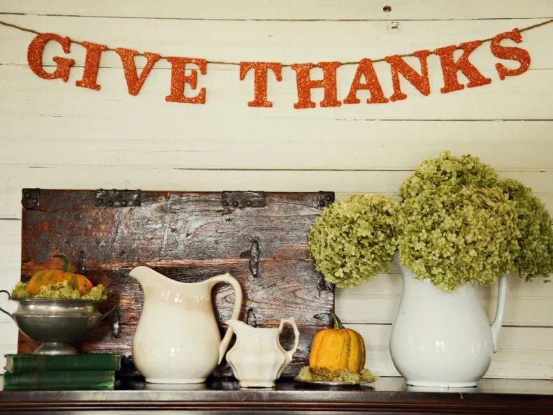 Orange Give Thanks letters hang above pale pitchers, small pumpkins and pale green flower heads on a dark surface