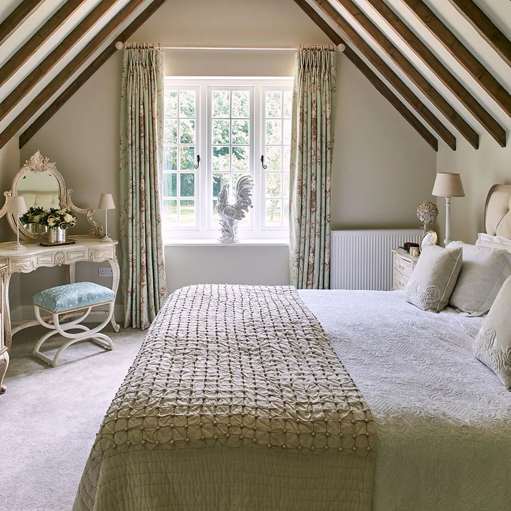 A pale bedroom sits beneath dark sloping beams, with patterned curtains, a cream bed, a small dressing table and an upholstered stool.