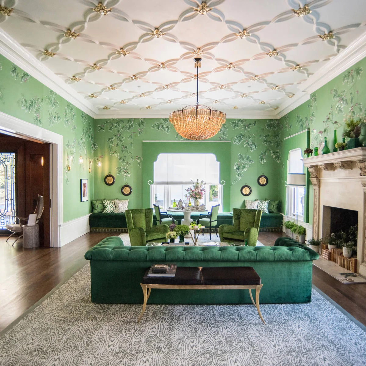 Rich green sofas and chairs sit on a broad gray patterned rug in a green-walled living room.