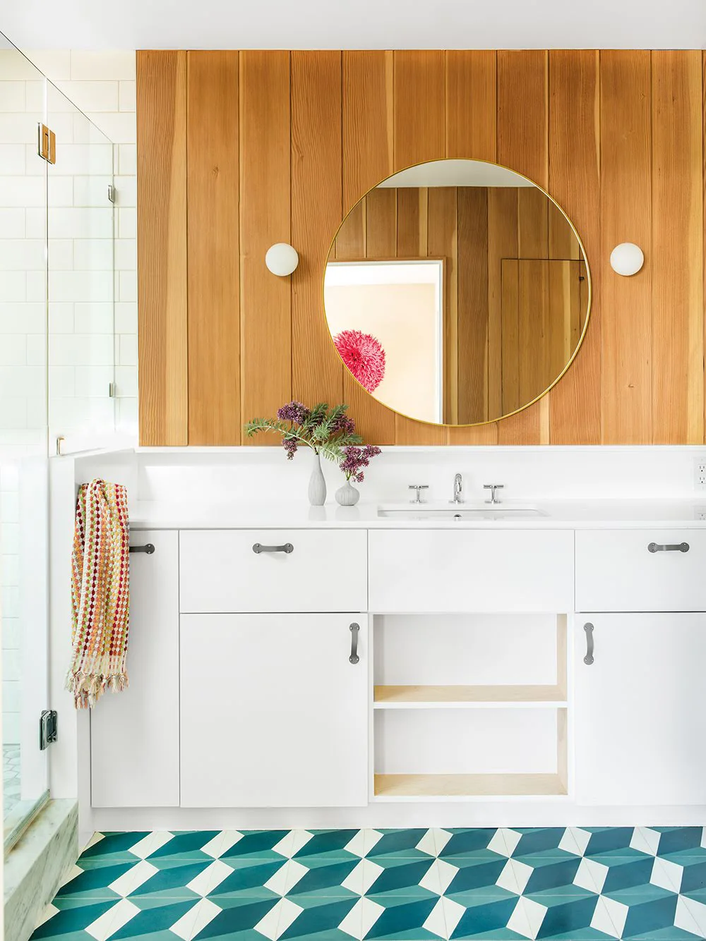 Vertical wood bathroom panels frame a large round mirror and two white globe lights above a pale vanity beside teal and white geometric floor tiles