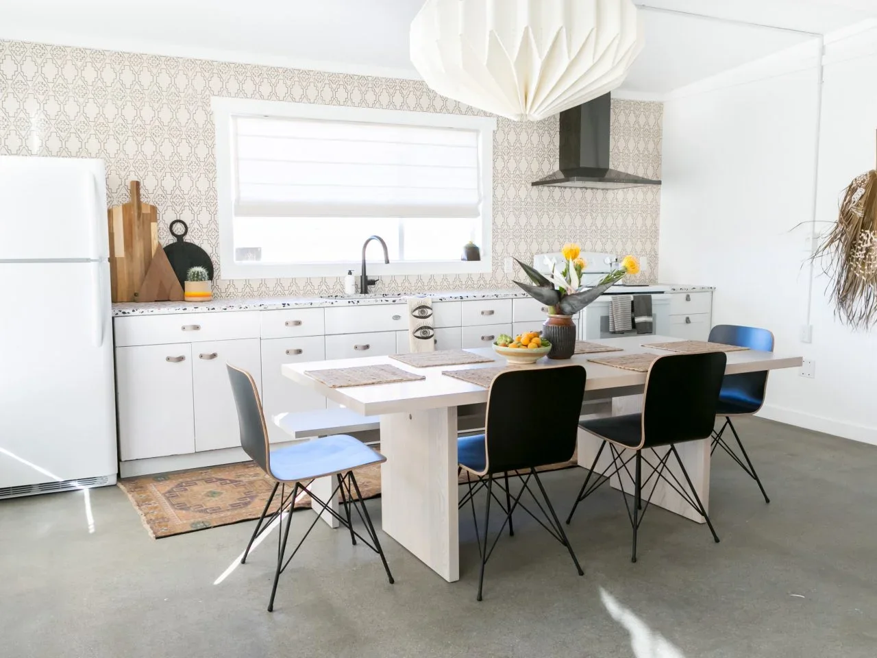 A compact white kitchen has a fine pale geometric backsplash around a window beside a small dining table.