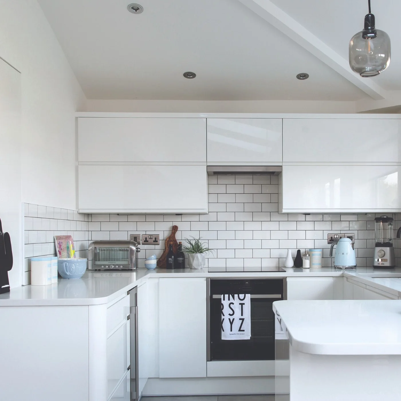 Glossy white cabinets with broad horizontal upper doors surround a compact U-shaped kitchen with white tiles and dark grout.