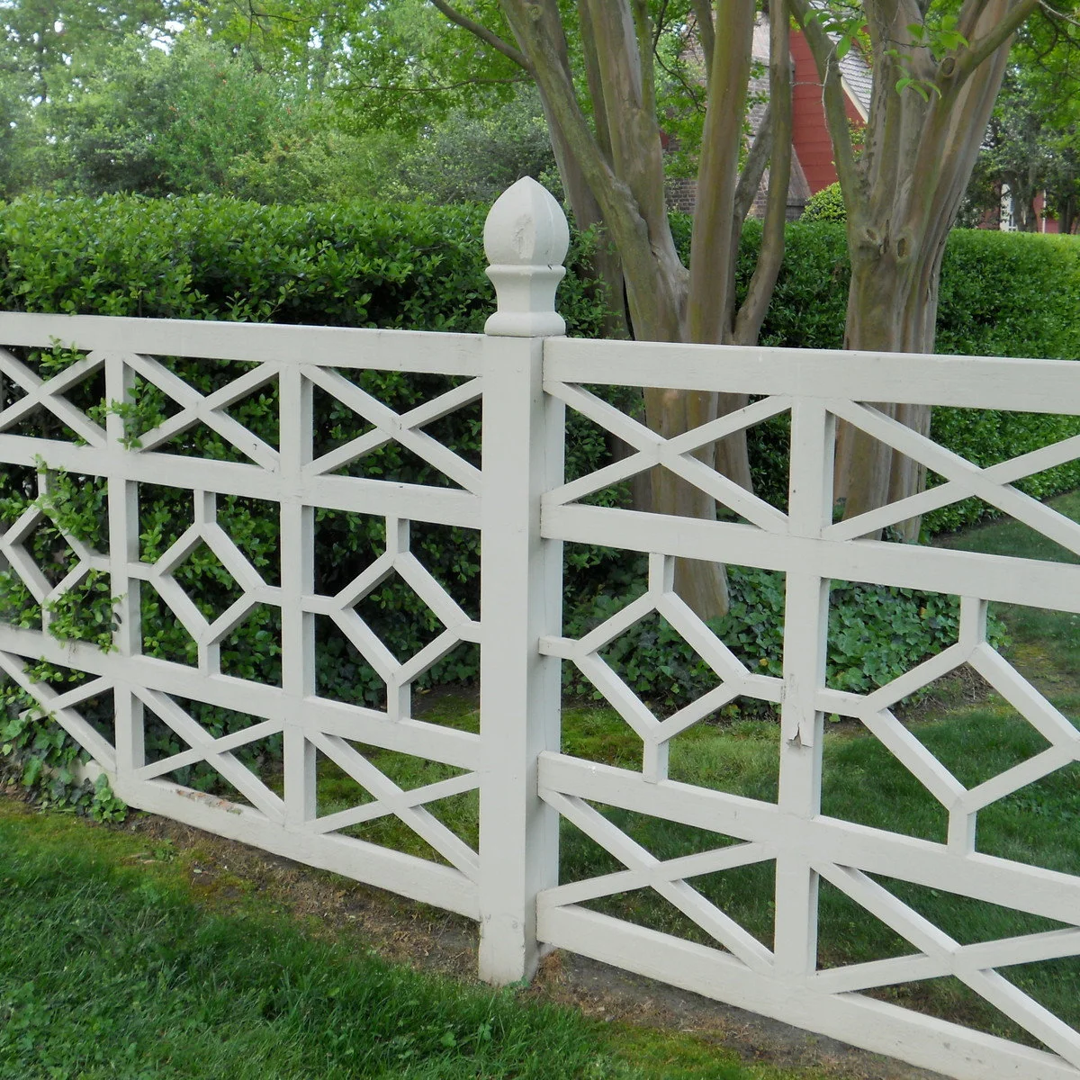 A white fence with diagonal crosses and central diamond shapes in front of a clipped hedge.