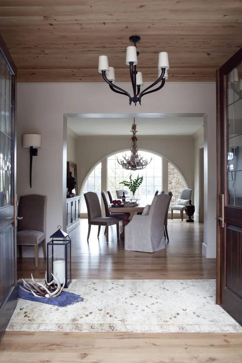 Dark curved chandelier with white shades under wood ceiling in entry opening toward dining room, with wall sconces, chair and pale rug